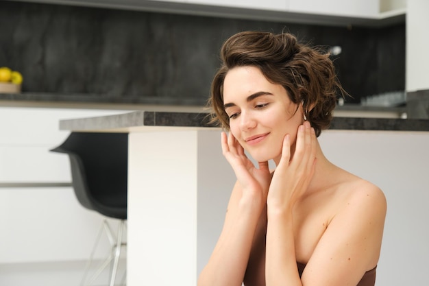 Photo vue rapprochée d'une jeune femme brune souriante. fille de fitness dans la cuisine. regarde de côté. touche son visage.