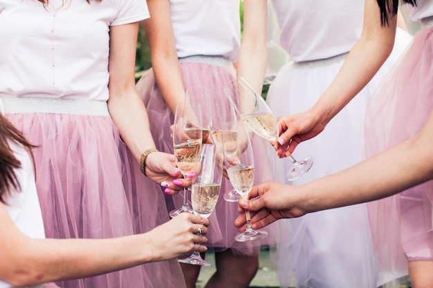 Vue rapprochée des mains des jeunes filles avec des verres de champagne. Photo recadrée de la mariée et de ses copines, toutes en jupes roses moelleuses, tenant des verres de vin à la main