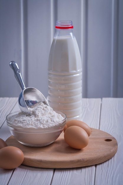 Vue rapprochée oeufs farine dans un bol scoop bouteille de lait sur une vieille planche de bois peinte en blanc concept de nourriture et de boisson