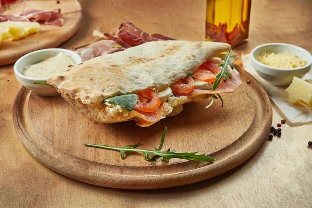 Photo vue rapprochée sur savoureux sandwich traditionnel italien panuozzo - tortilla cuite au four avec des tomates, de la mozzarella, du jambon et de la roquette sur une planche de bois. l'alimentation de rue