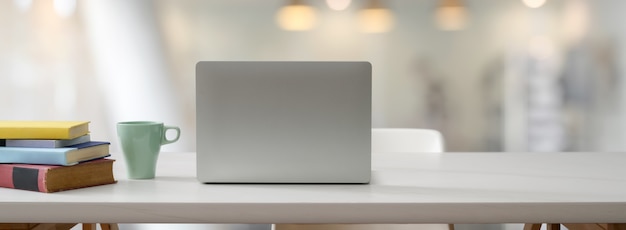 Vue rapprochée d'une table de travail confortable avec un ordinateur portable ouvert, des livres, une tasse et un espace de copie sur un tableau blanc