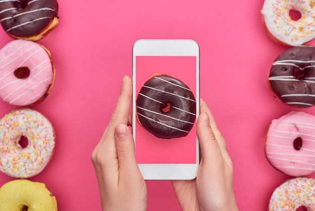 Vue recadrée d'une femme tenant un smartphone avec une image de beignet sur fond rose près de délicieux beignets