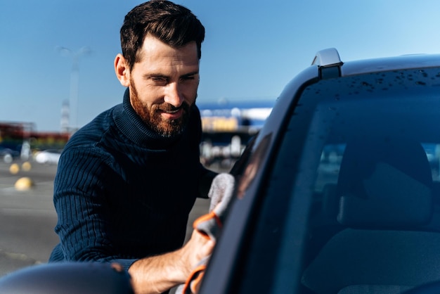 Vue recadrée de l'homme barbu avec une lingette en microfibre grise polissant sa nouvelle voiture électrique Bel homme caucasien lavant et essuyant sa voiture avec plaisir sourire Concept de lavage de voiture