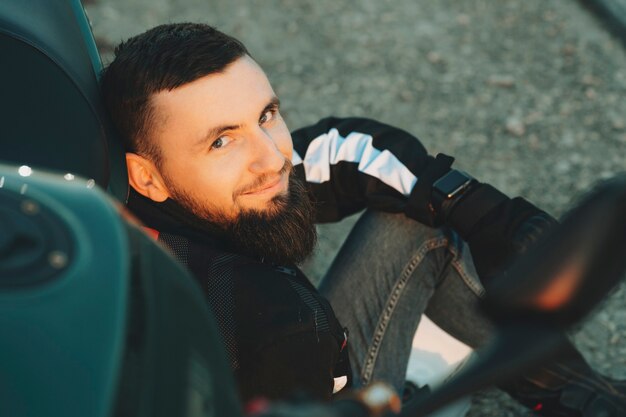 Vue de la récolte de l'heureux jeune homme barbu en veste de motard assis sur la route se penchant à moto en regardant la caméra