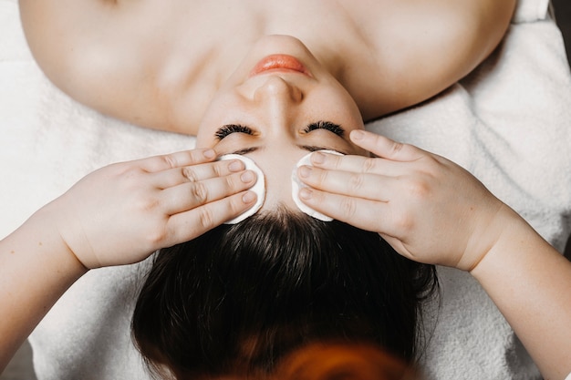 Vue supérieure d'une main de cosmétologue faisant la routine de soins de la peau avant le traitement du visage sur un joli visage féminin dans un centre de bien-être.