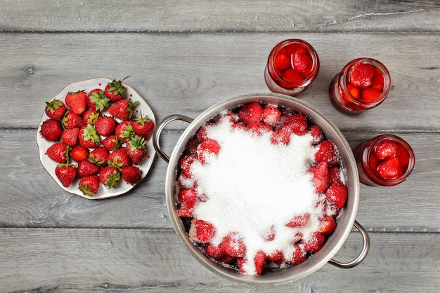 Vue de table, grand pot en acier avec des fraises recouvertes de sucre cristal, trois flacons avec des fruits au sirop et une assiette à côté. Préparation de compote de fraises maison.