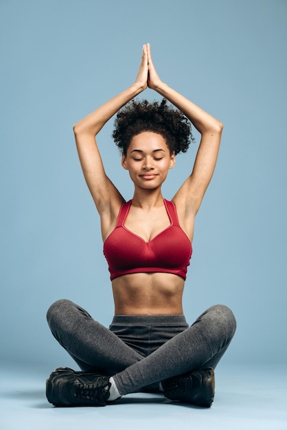 Vue sur toute la longueur de la femme consciente faisant un geste de prière assise en position du lotus dans le studio aux murs bleus. Stock photo