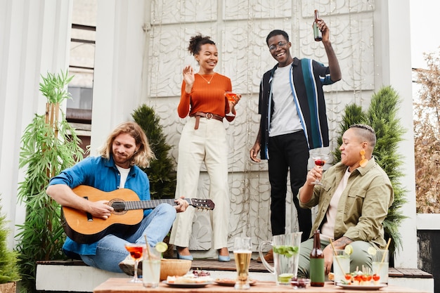 Vue sur toute la longueur d'un groupe d'amis dansant lors d'une fête en plein air sur le toit, avec un jeune homme jouant de la guitare au premier plan, espace pour copie