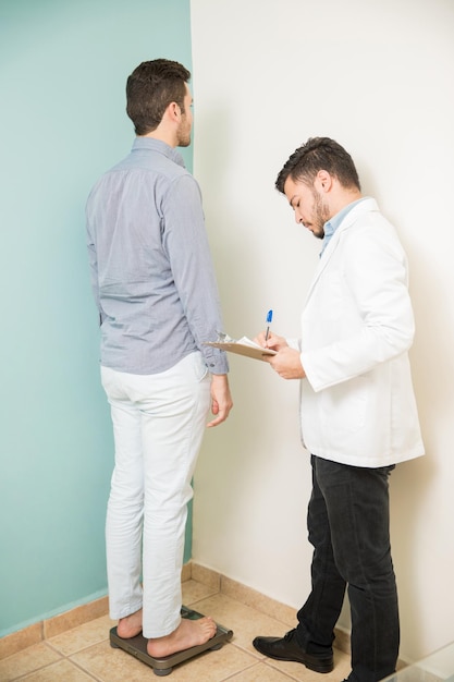 Vue sur toute la longueur d'un nutritionniste masculin prenant des notes pendant qu'un patient se tient sur une balance dans une clinique de santé