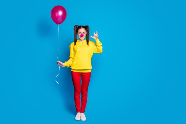 Vue sur toute la longueur de la taille du corps d'une jolie fille effrayante et méchante folle agressive clown de cirque tenant dans la main une balle d'hélium faisant semblant d'attaquer isolée sur fond de couleur bleu vif éclatant brillant