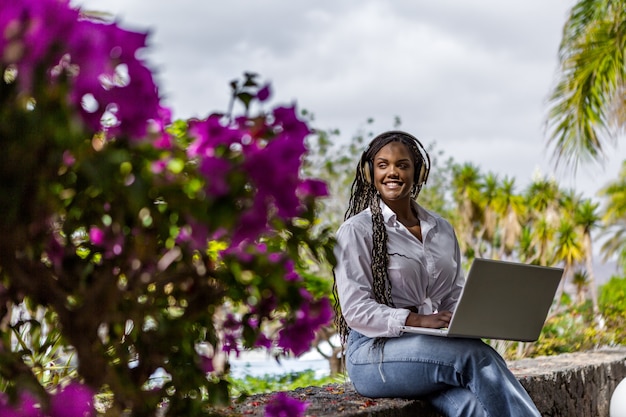 Vue à travers une fleur violette d'une femme se relaxant à l'extérieur à l'aide d'un ordinateur portable tout en écoutant de la musique en détournant les yeux. Femme afro-américaine profitant de la journée.