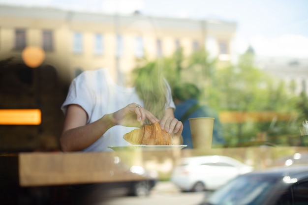 Une vue à travers le verre d'une fille dans un café qui boit un verre et mange un croissant
