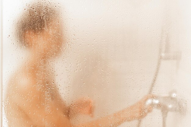 Vue à travers la vitre d'une cabine de douche d'une femme caucasienne méconnaissable prenant une douche dans un hôtel avec espace de copie.