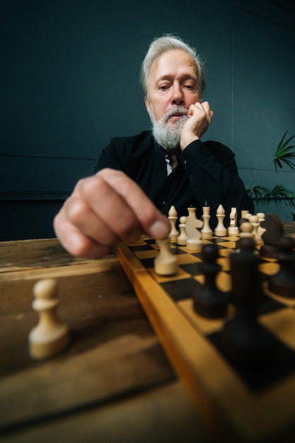 Vue verticale en angle bas d'un homme mature aux cheveux gris concentré effectuant un mouvement avec une pièce de pion sur un échiquier en bois, mise au point sélective. Bel homme barbu jouant aux échecs seul à la maison.