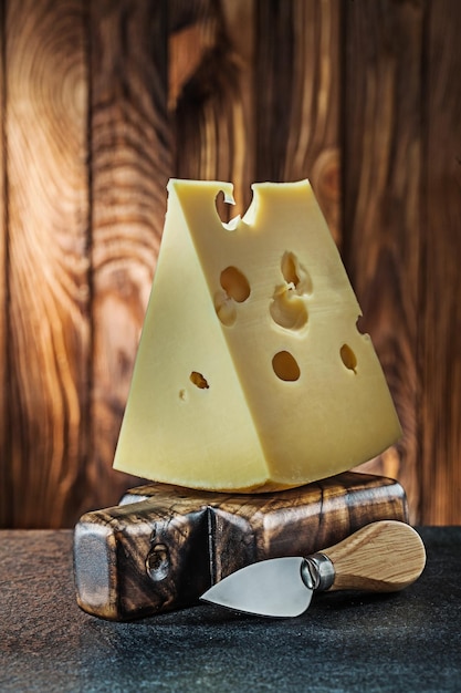 Vue verticale gros morceau de lait fromage maasdam sur petite planche à découper et couteau vieux fond en bois