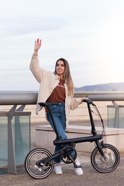 Vue verticale d'une jeune femme saluant quelqu'un tout en tenant un vélo électrique pour rentrer à la maison depuis le centre-ville