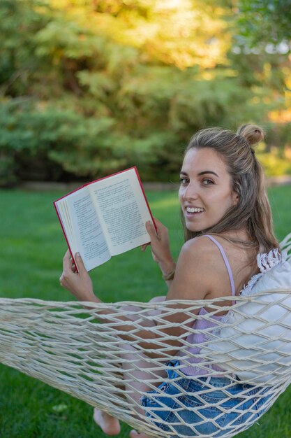 Vue verticale d'une jeune femme souriante regardant la caméra tout en lisant un livre dans un hamac dans le jardin en été