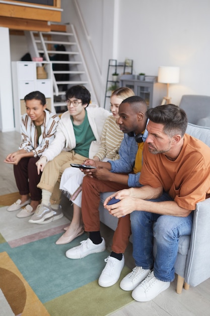 Vue verticale pleine longueur à divers groupes d'amis regardant la télévision à la maison ensemble tout en étant assis en rang sur un canapé