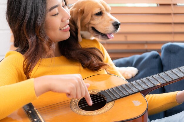 Un week-end Une jeune femme asiatique et son chiot beagle jouent de la guitare et chantent joyeusement ensemble dans le salon d'une maison