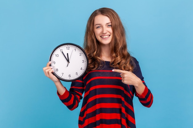 Winsome femme portant un pull rayé de style décontracté, pointant une grande horloge et regardant la caméra avec positif, montrant des montres murales pour se dépêcher. Studio intérieur tourné isolé sur fond bleu.