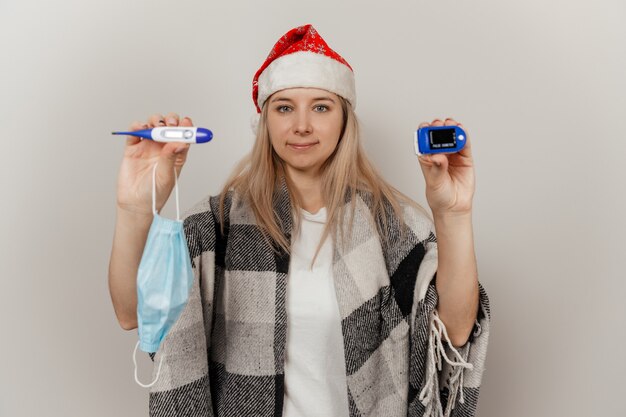 Woman in santa hat and wrap détient thermomètre et oxymètre de pouls sur fond isolé gris