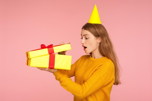 Wow beau cadeau Portrait d'une fille au gingembre étonnée avec un cône de fête sur la tête regardant à l'intérieur d'une boîte-cadeau avec intérêt et étonnement déballant une surprise d'anniversaire studio intérieur tourné sur fond rose