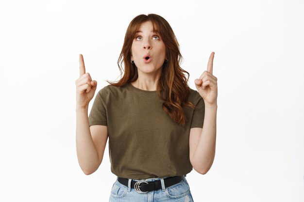 Wow génial. Une fille impressionnée lève la tête, pointant du doigt la publicité du haut avec une expression de visage fascinée debout sur blanc.