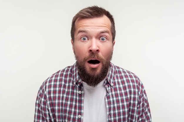 Wow incroyable Portrait d'un homme barbu effrayé choqué en chemise à carreaux décontractée regardant la caméra avec de grands yeux étonnés sa bouche s'ouvrit en studio intérieur surprise tourné isolé sur fond blanc