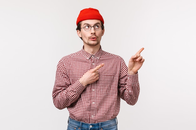 Wow look intéressant là-bas. Intrigué jeune homme de race blanche avec barbe en bonnet rouge et lunettes, furtivement pointant le coin supérieur droit avec intérêt et curiosité,