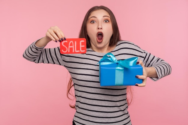 Wow, des réductions d'anniversaire. Portrait d'une femme surprise en sweat-shirt rayé tenant l'inscription Vente, boîte-cadeau et regardant la caméra avec un visage étonné choqué. tourné en studio isolé sur fond rose