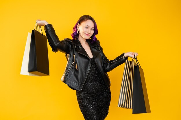 Wow shopping femme heureuse vendredi noir vente de saison rabais festif dame élégante veste en cuir cheveux bouclés violet montrant des sacs d'achat jaune isolé