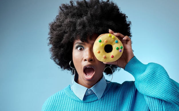 Photo wow surprise et beignet sur l'œil d'une femme noire se sentant choquée par un dessert au sucre ou des bonbons sur fond bleu studio bonbons et gâteau avec fille et omg émotion pour vente ou alerte