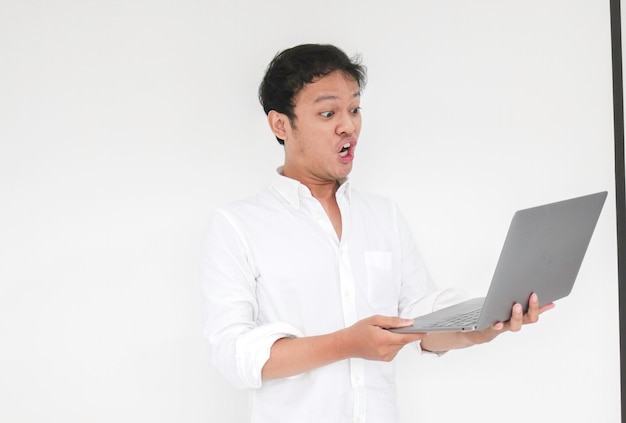 Wow visage de jeune homme asiatique choqué ce qu'il voit dans l'ordinateur portable lorsqu'il travaille sur fond gris isolé portant une chemise blanche