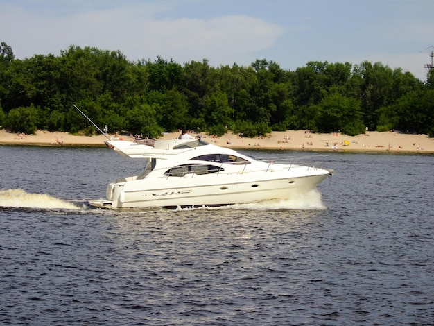 Un yacht blanc à grande vitesse navigue sur le fond de la plage avec des gens et des arbres en train de bronzer