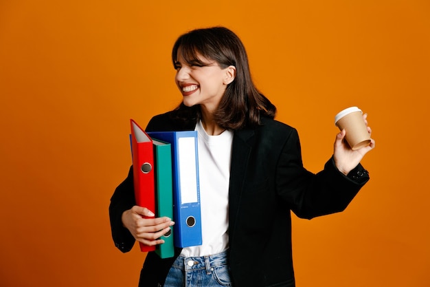 Les yeux fermés tenant des dossiers avec une tasse de café belle jeune femme portant une veste noire isolée sur fond orange