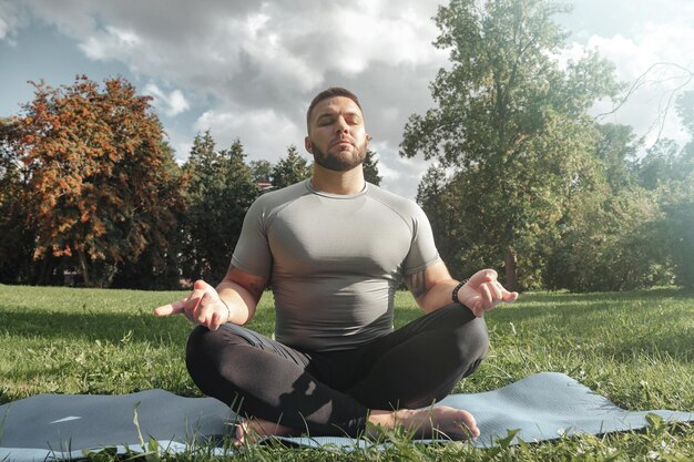 Yogi de jeune homme sportif assis dans la pose de yoga de lotus dans le parc de pelouse d'herbe à l'extérieur