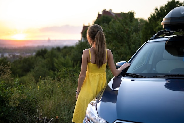 Yong happy conductrice se reposant près de sa voiture en profitant de la vue du coucher de soleil sur la nature estivale Concept de voyage et de loisirs