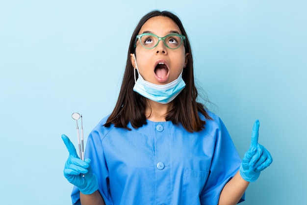 Young brunette mixed race dentist woman holding tools sur fond isolé surpris et pointant vers le haut