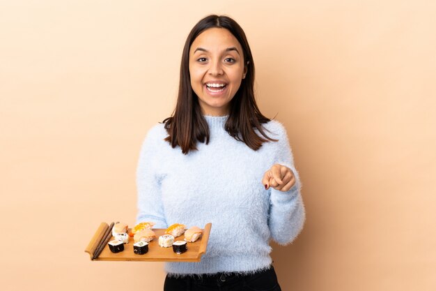 Young brunette mixed race woman holding sushi sur isolé surpris et pointant vers l'avant