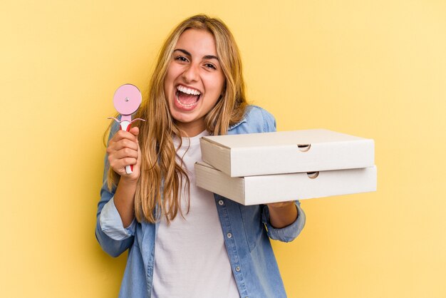 Young caucasian blonde woman holding pizzas et cutter isolé sur fond jaune