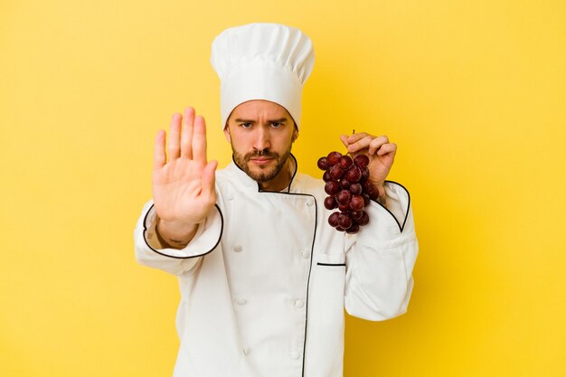 Young caucasian chef man holding raisins isolés sur fond jaune debout avec la main tendue montrant le panneau d'arrêt, vous empêchant.