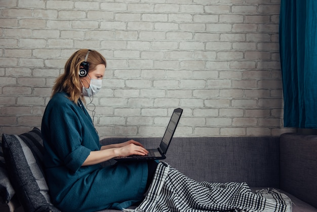 Young caucasian European woman wearing medical mask et plaid à l'aide d'un ordinateur portable sur le canapé pendant le travail à distance à la maison en raison de la pandémie de coronavirus