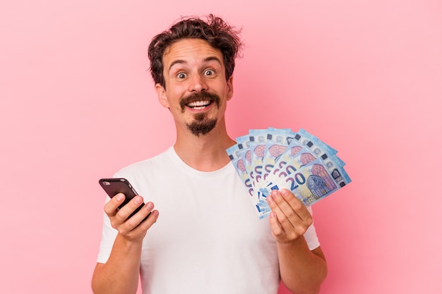 Young caucasian man holding bills et tirelire isolé sur fond rose