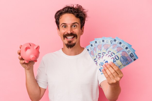 Young caucasian man holding bills et tirelire isolé sur fond rose