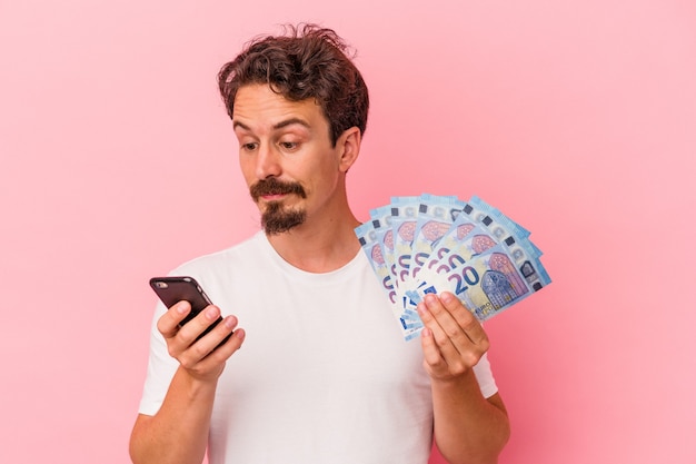 Young caucasian man holding bills et tirelire isolé sur fond rose