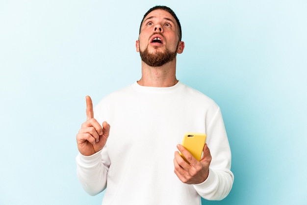 Young caucasian man holding mobile phone isolé sur fond bleu pointant vers le haut avec la bouche ouverte.