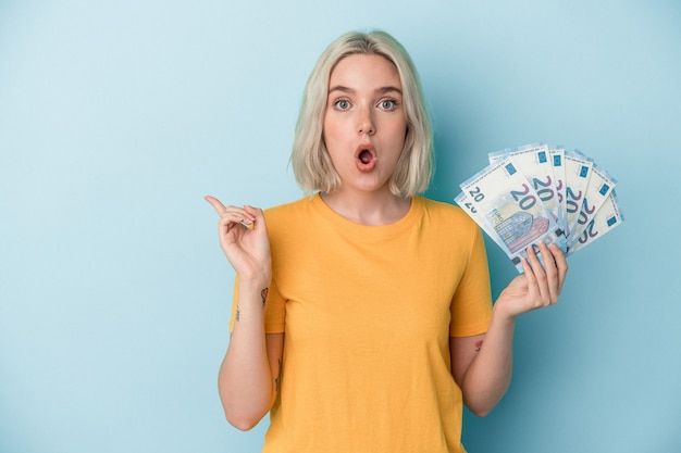 Young caucasian woman holding bills isolé sur fond bleu pointant vers le côté