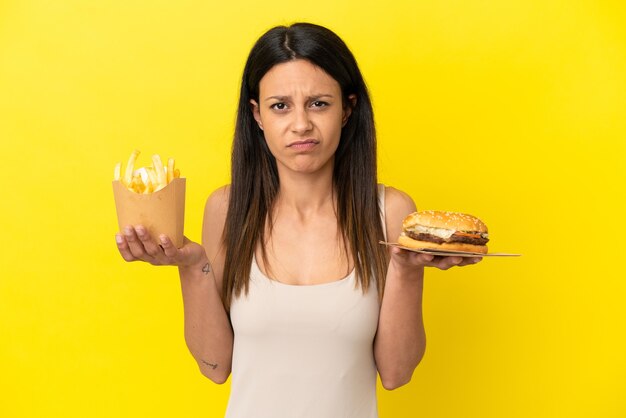 Young caucasian woman holding burger et chips isolé sur fond jaune