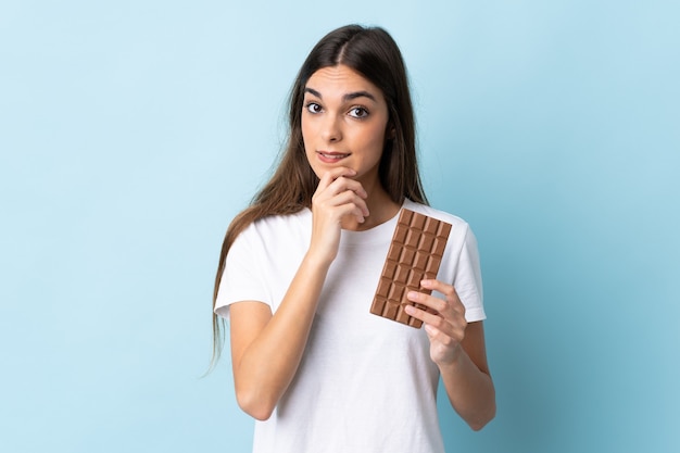 Young caucasian woman isolated on blue prenant une tablette de chocolat et ayant des doutes