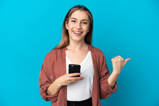 Young caucasian woman isolated on blue wall using mobile phone et pointant vers le côté